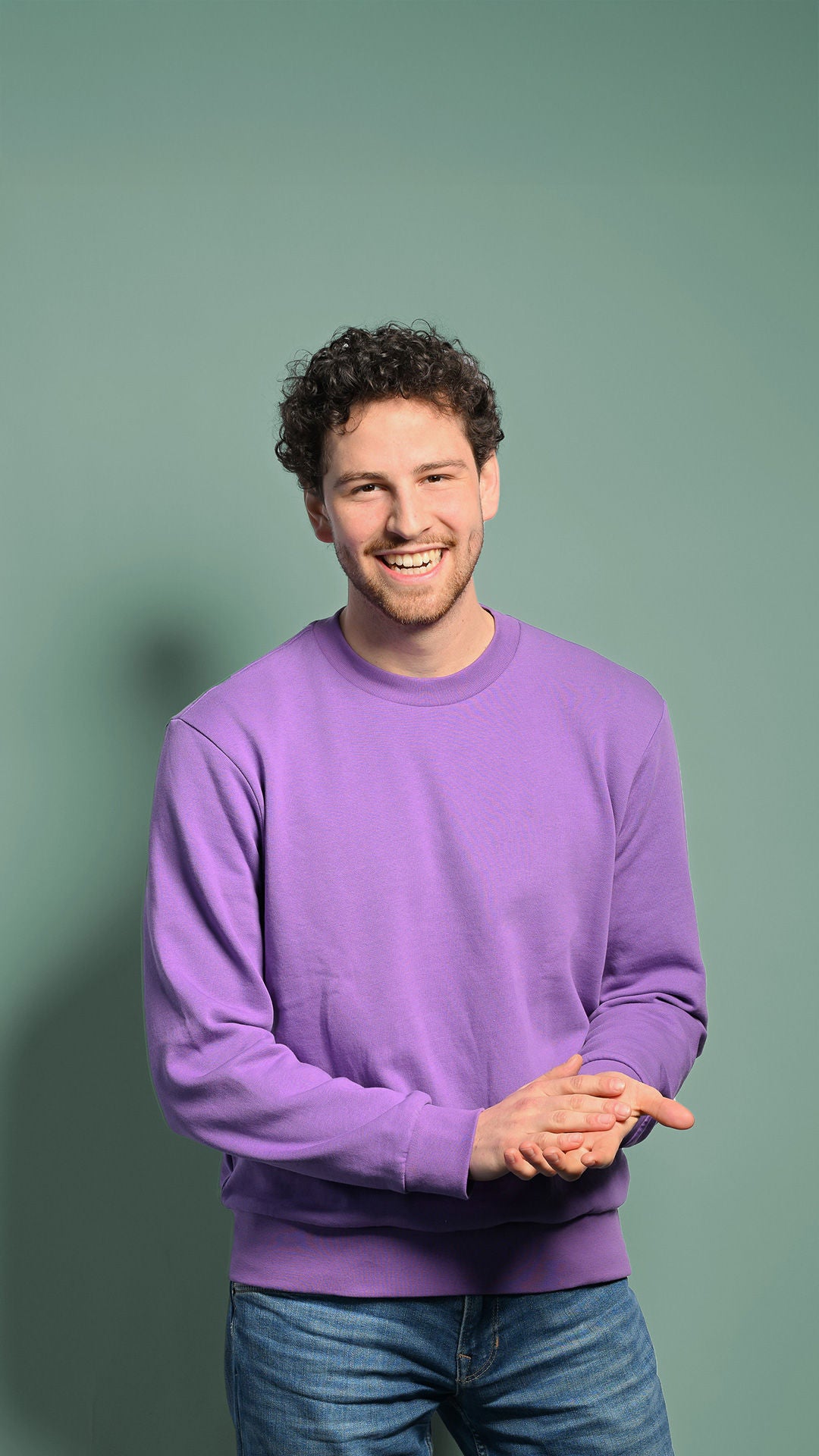 Fabian steht mit einem lila Pullover vor einer Wand und lacht an der Kamera vorbei