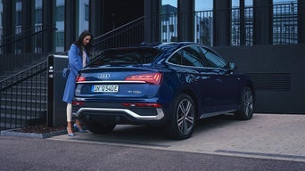 Frau in blauem Mantel steht neben einem blauen Audi Q5 e-tron an einer Ladestation vor einem modernen Gebäude.