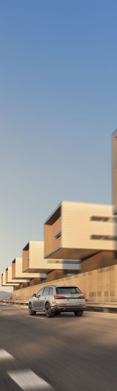 Rückansicht eines fahrenden, silbernen Audi Q7 SUV TFSI e auf leerer Straße, modernes Architekturensemble im Hintergrund.