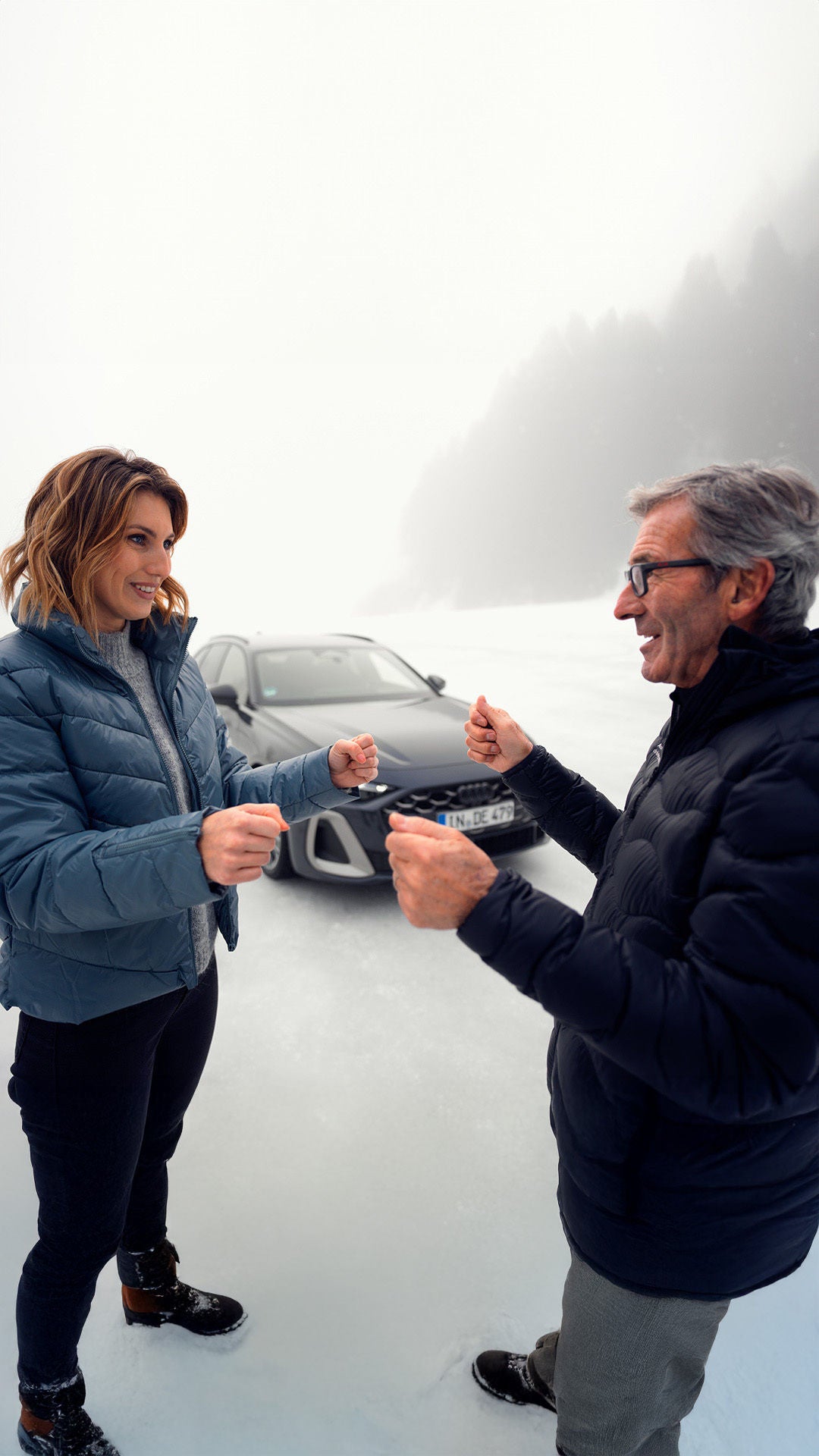 Zwei Personen (jüngere Frau und älterer Mann mit Brille) in Winterkleidung stehen auf einer Eisfläche und simulieren Fahrbewegungen, im Hintergrund ein Audi Fahrzeug.