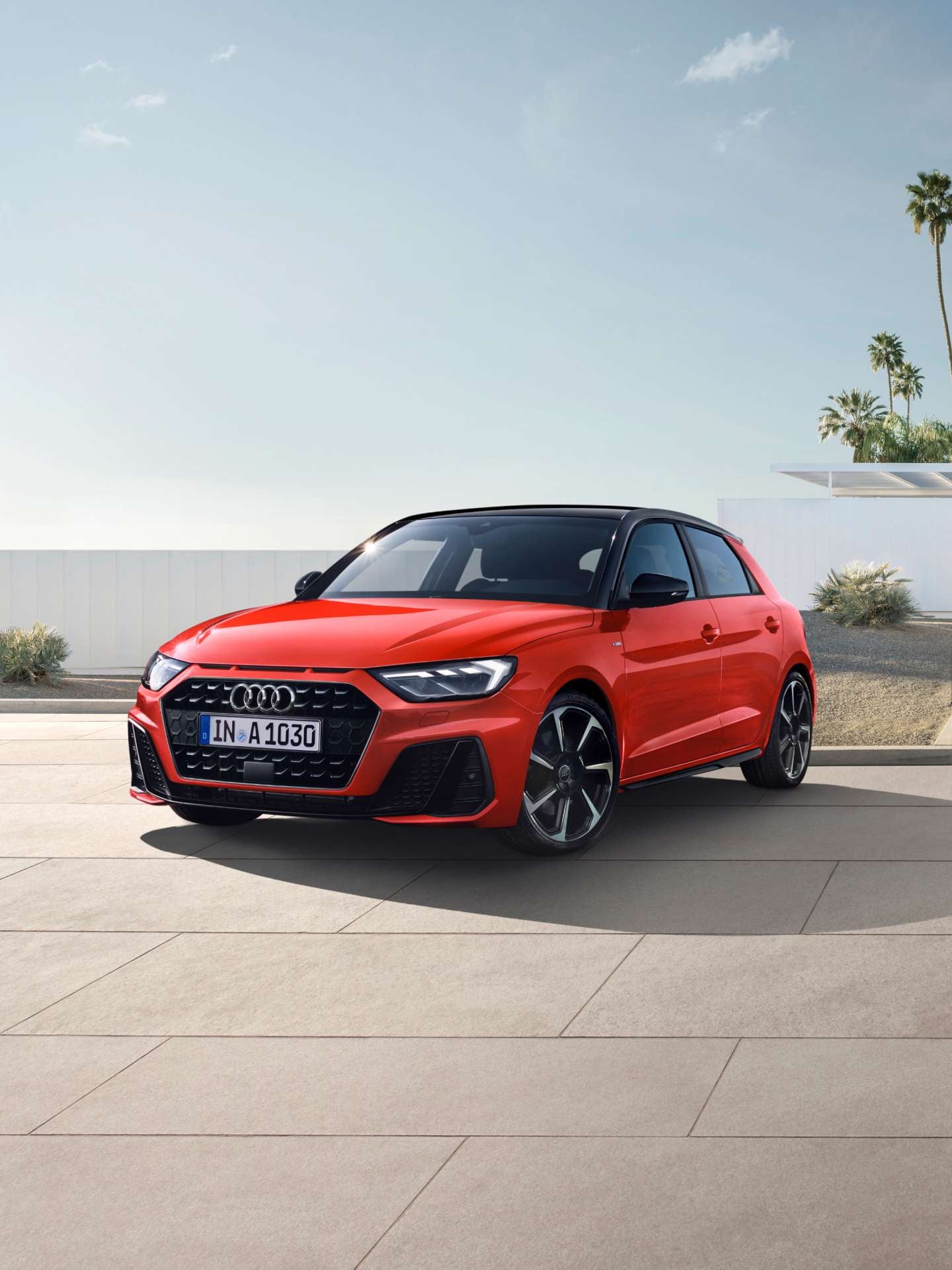 A red Audi A1 Sportback car parked on a paved road near a modern building with palm trees in the background.