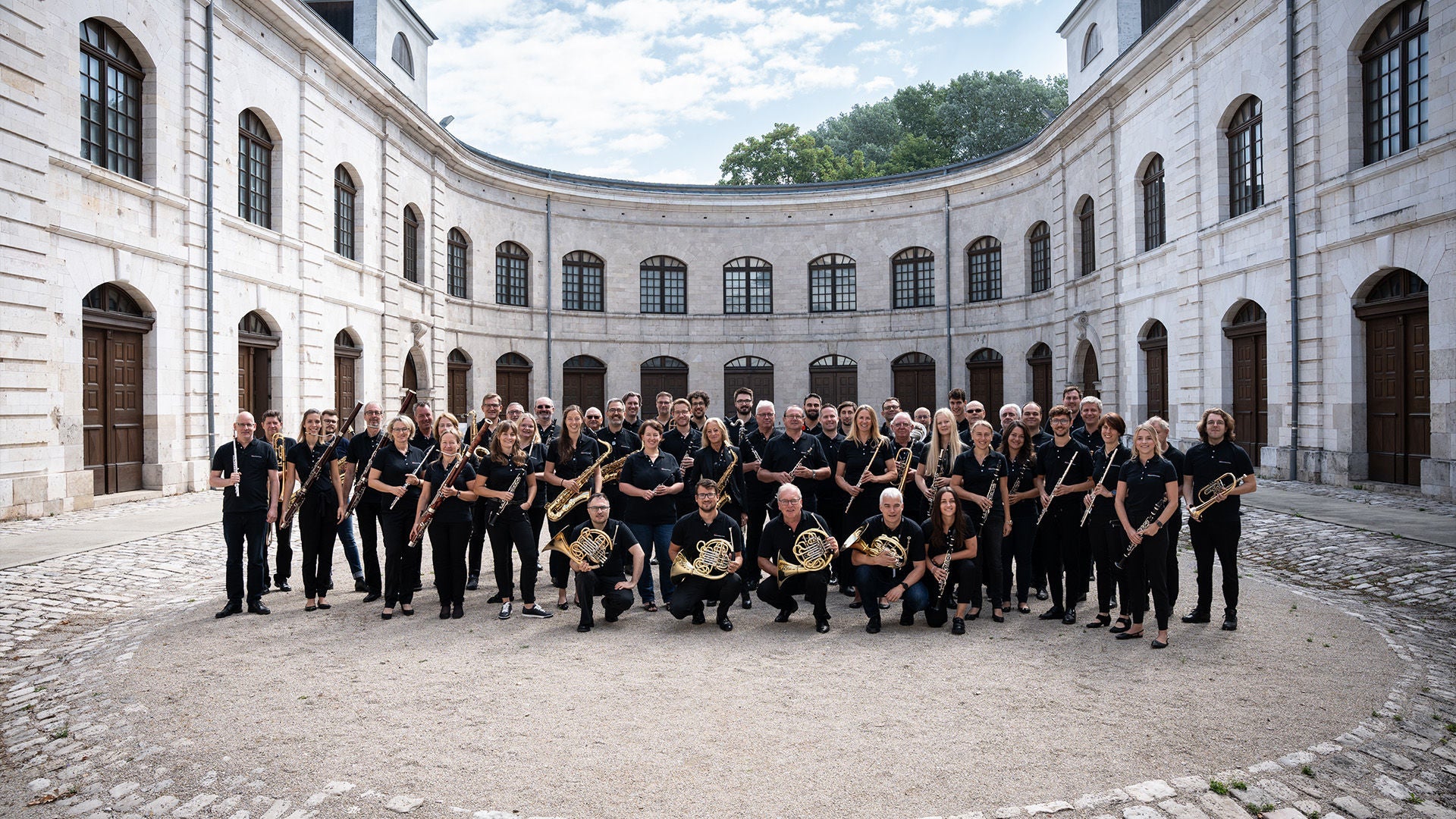 Großes Blasorchester in schwarzer Kleidung mit Instrumenten in einem hellen, halbkreisförmigen Innenhof.