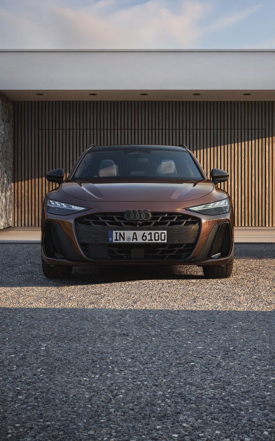 Brauner Audi A6 Avant Frontansicht, geparkt vor moderner Architektur mit Holz- und Steinfassade bei sonnigem Wetter.