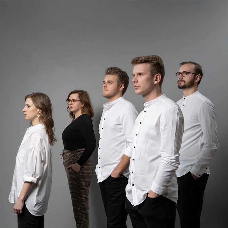 Fünf junge Erwachsene stehen seitlich nebeneinander vor grauem Hintergrund, drei in weißen Hemden, zwei in dunkler Kleidung.