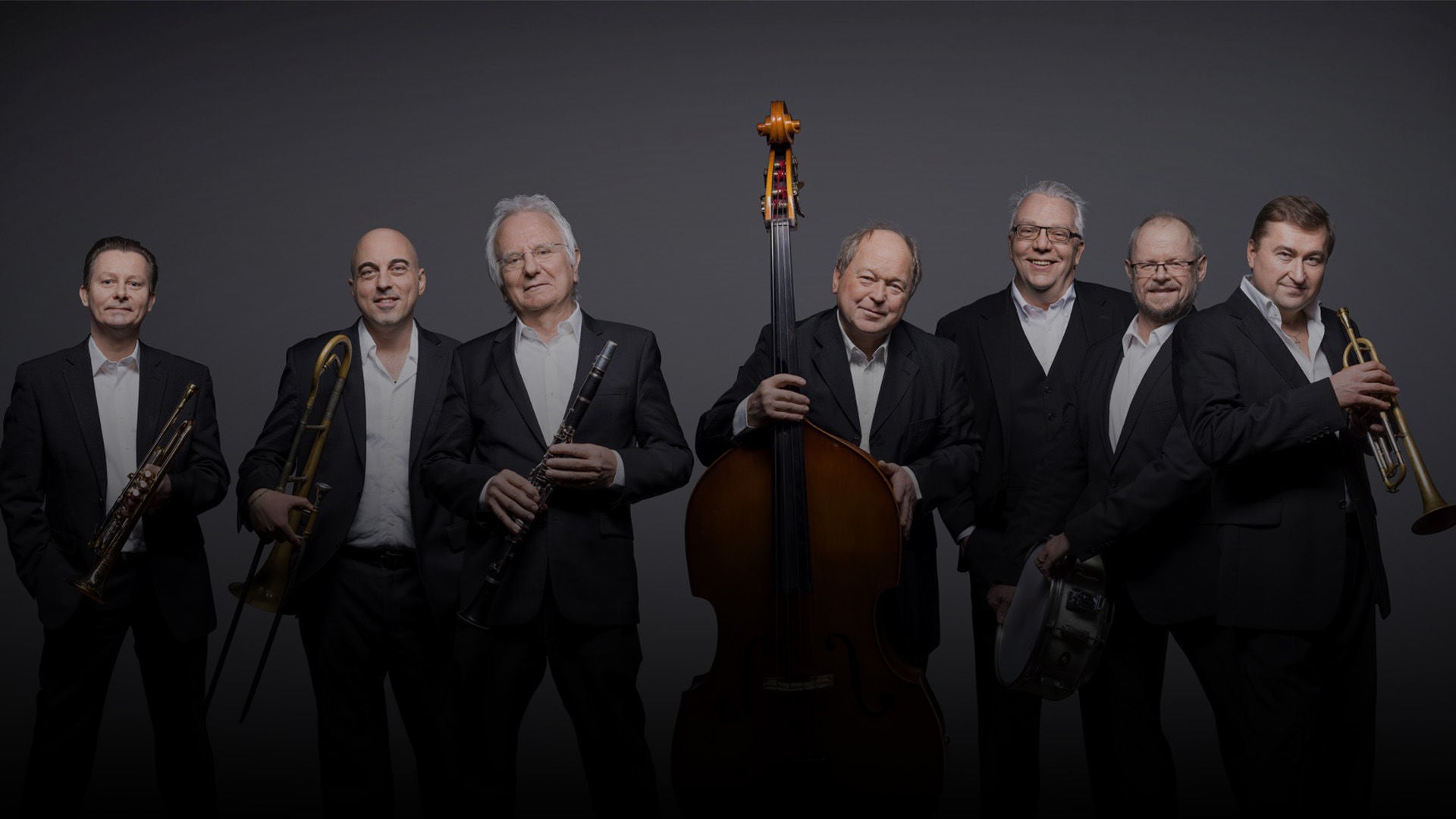 Sieben Jazzmusiker in dunklen Anzügen posieren vor grauem Hintergrund mit Trompeten, Posaune, Klarinette, Kontrabass und Schlagzeug.