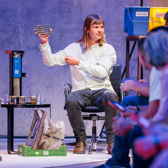 Ein Mann mit langen Haaren zeigt dem Publikum ein rechteckiges Objekt, im Hintergrund Recycling-Boxen.