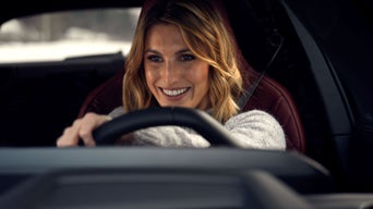 A smiling woman behind the wheel of an Audi with red seats, wearing her seatbelt; the interior is dark, and the background is out of focus.