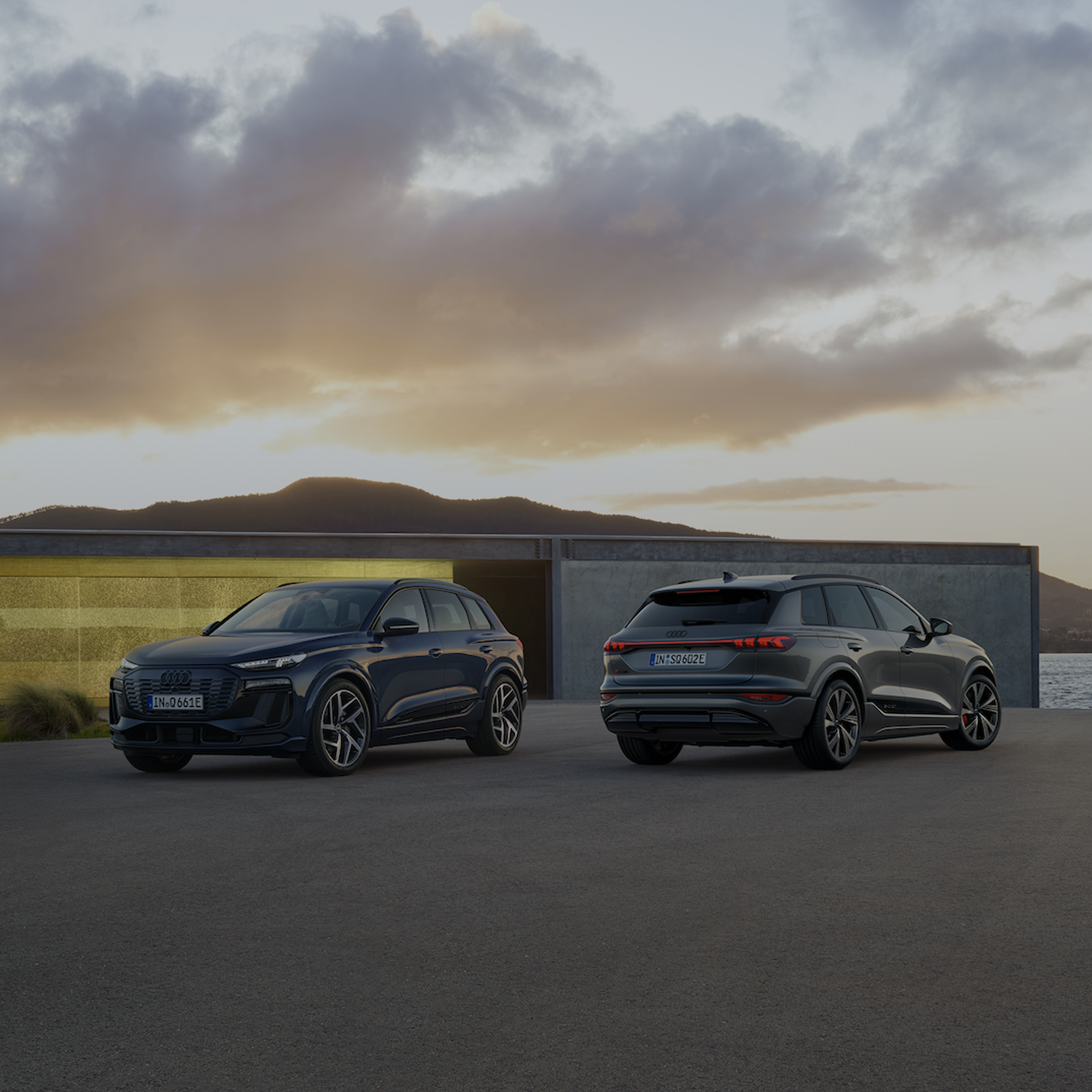 Two Audi cars side by side, one facing away with a yellow wall and a body of water in the background