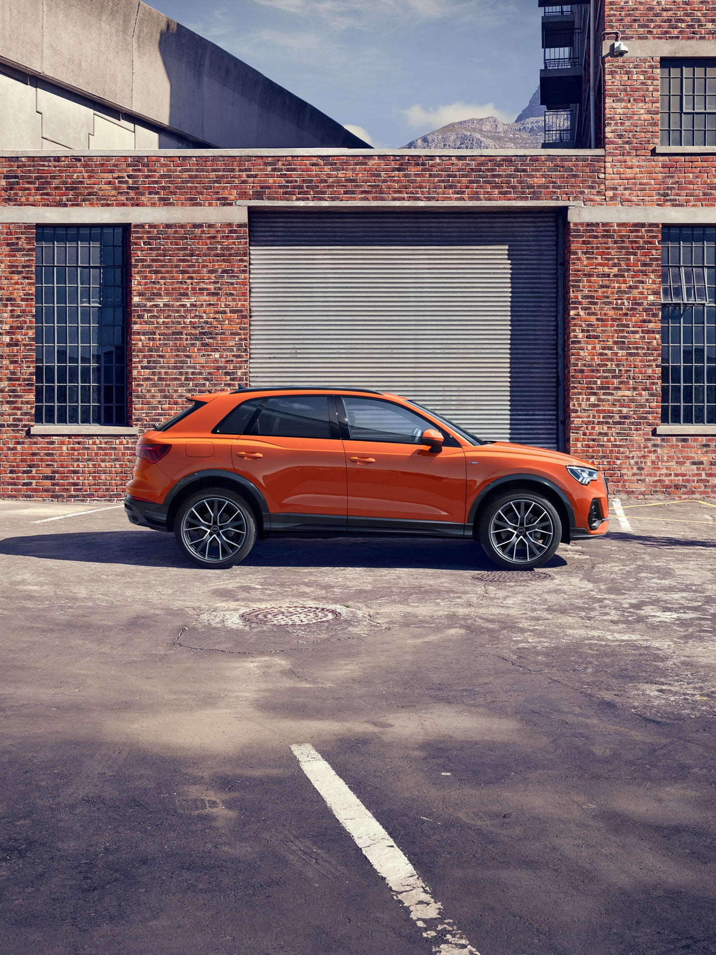Oranger Audi Q3 SUV parkt auf einem leeren, urbanen Hof vor einer Backsteinwand mit geschlossenem Rolltor und Fenstern.
