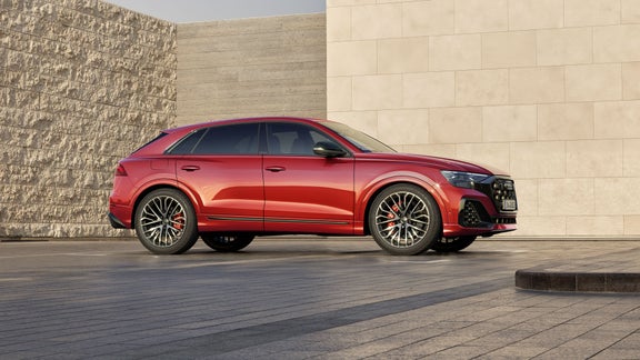Roter Audi SQ8 SUV parkt vor moderner Steinfassade bei Tageslicht, Seitenansicht mit sportlichem Design.
