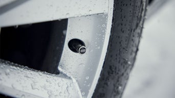 Nahaufnahme eines Autoreifens mit silberner Ventilkappe mit Audi-Ringen, benetzt mit Wassertropfen.