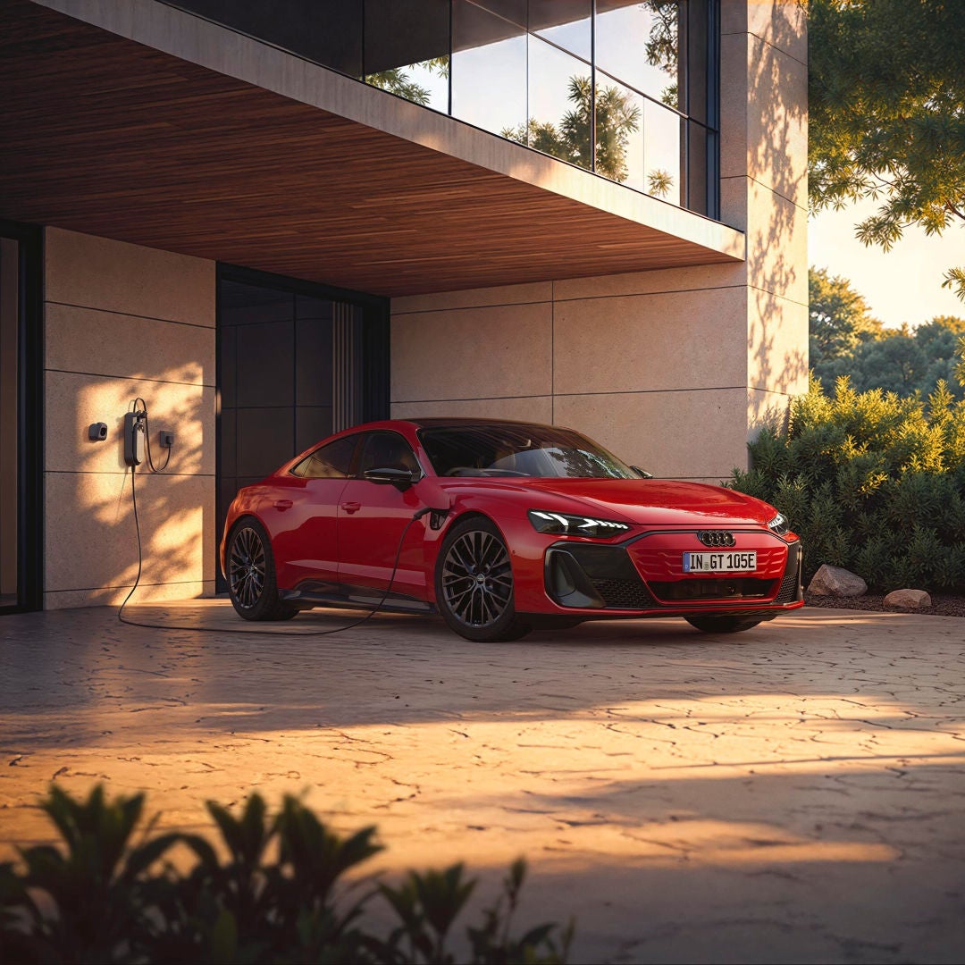 A sleek red electric Audi sports car is plugged in at home, set against a modern architectural backdrop with greenery.
