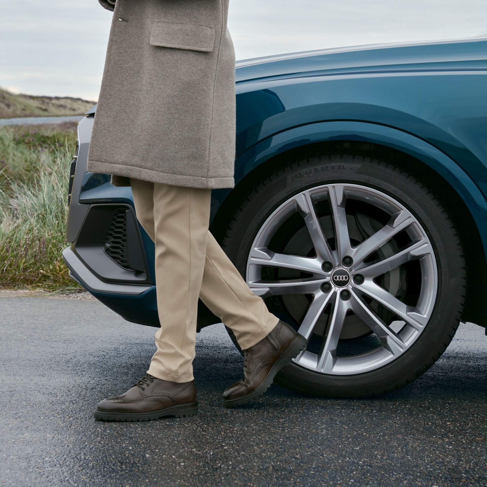 Person in beigem Mantel steht neben einem blauen Audi mit silberner Alufelge auf nasser Straße.