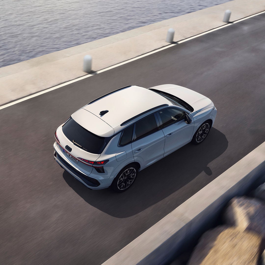 A white Audi Q3 Sportback e-hybrid SUV drives on a coastal road beside calm water, viewed from above in bright daylight.
