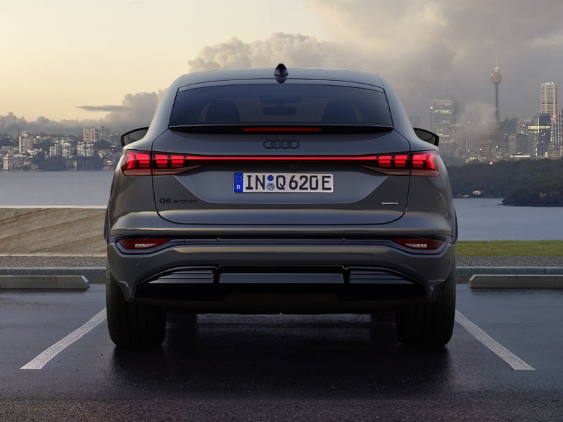 Heckansicht des Q6 Sportback e-tron, welcher auf einem Parkplatz am Meer steht. Im Hintergrund sieht man eine Stadt, welche von Wolken durchzogen ist.