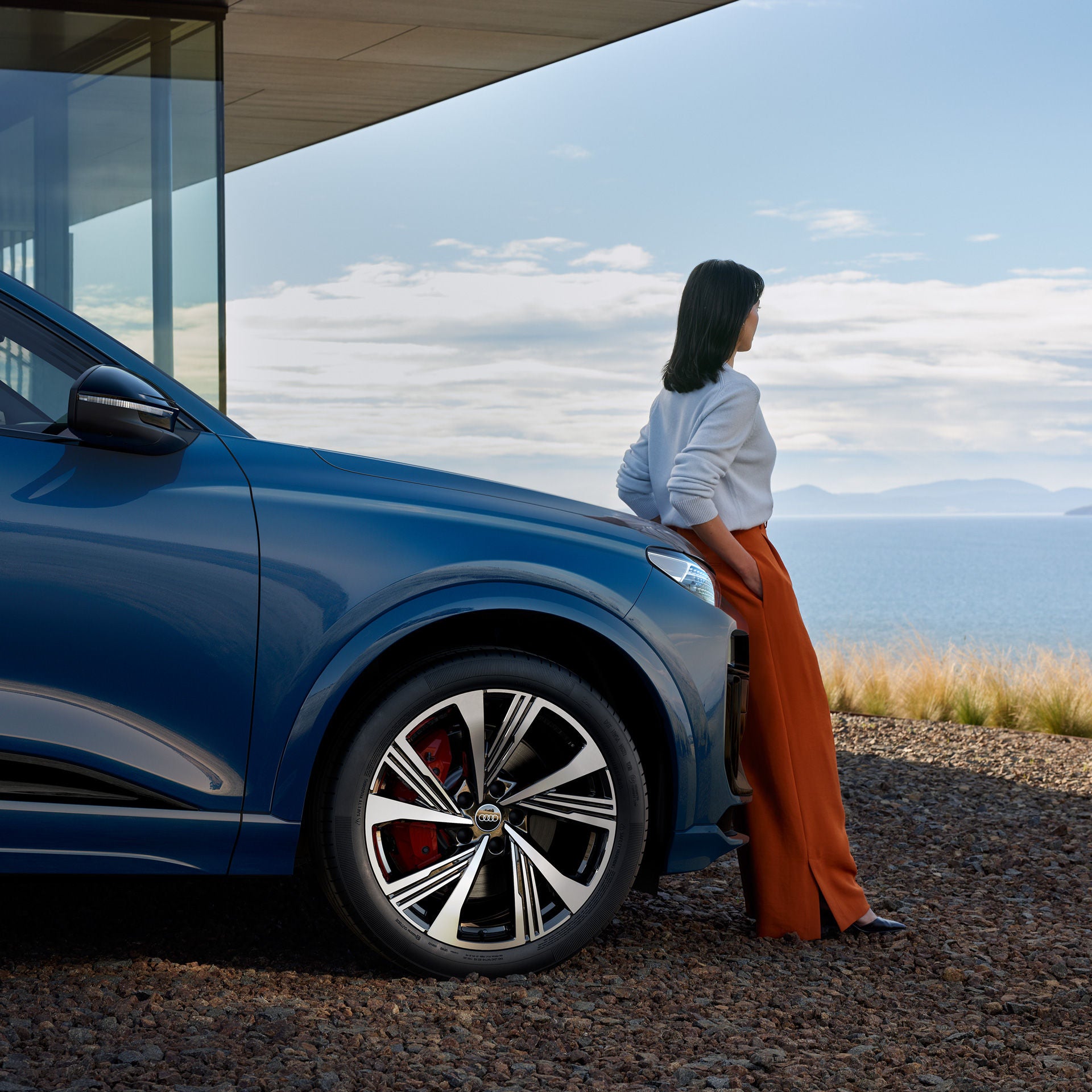 Frau lehnt an einem blauen Audi SUV mit Blick aufs Wasser, im Hintergrund Berge und blauer Himmel mit Wolken.