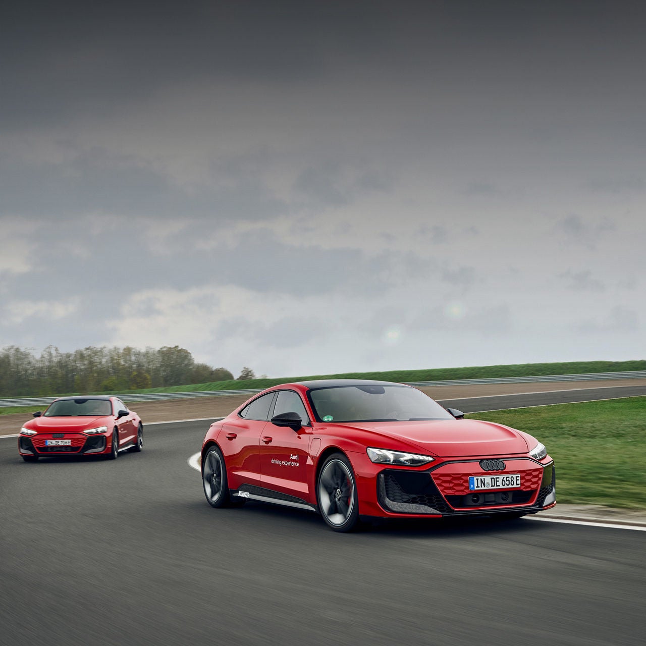 Zwei rote Audi e-tron GT fahren dynamisch auf einer Rennstrecke bei bewölktem Himmel.