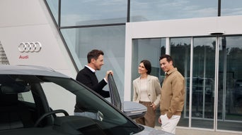Drei Personen stehen vor Audi-Autohaus, ein Mann öffnet die Tür eines grauen Fahrzeugs und spricht mit einem Paar.