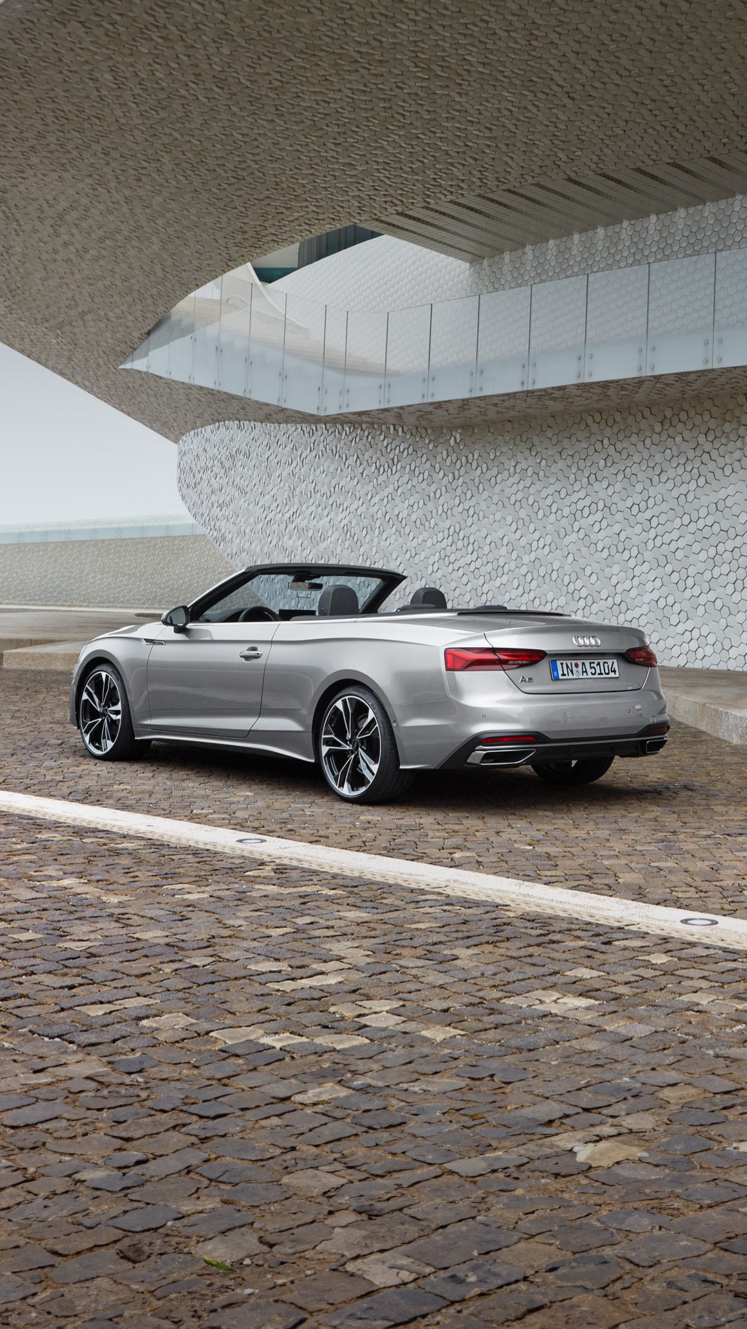Silbernes Audi A5 Cabriolet mit offenem Verdeck steht vor moderner Architektur mit heller Mosaikwand auf Kopfsteinpflaster.