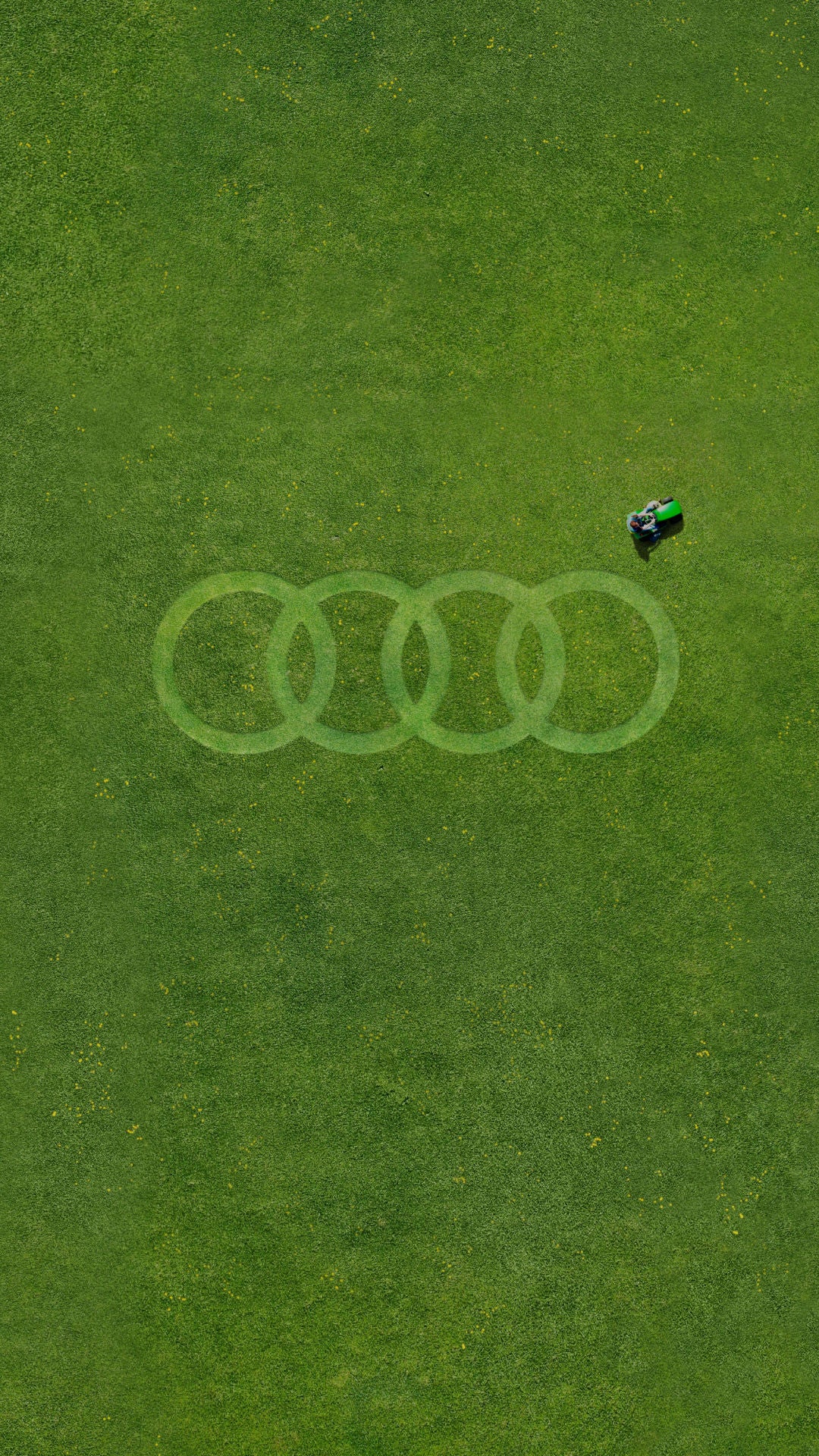 Aerial view of a lush green lawn with a person mowing, creating the Audi logo pattern in the grass.