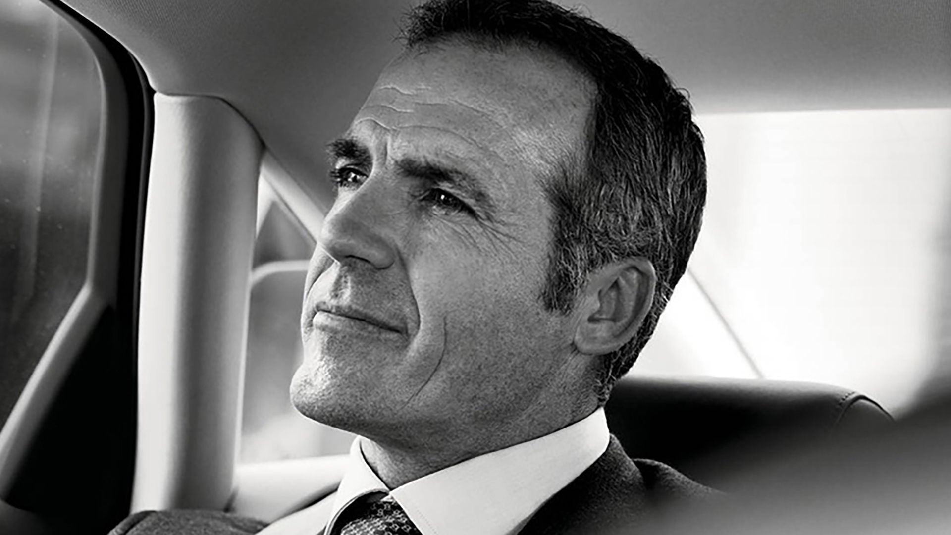 Black and white portrait of a man in a suit sitting relaxed in the back seat of a car, looking out of the window