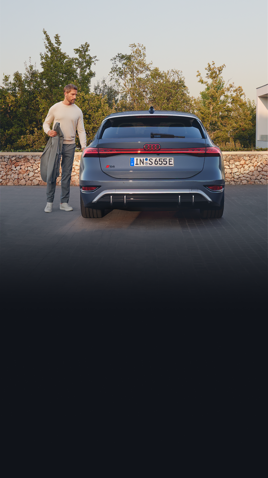 Rear view of the S6 Avant e-tron parked on cobblestones in front of a stone wall and the garden of a modern house. A middle-aged man is about to open a door.