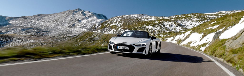 White Audi R8 Spyder V10 performance quattro drives with the top down under blue skies on a winding road through the Alps.