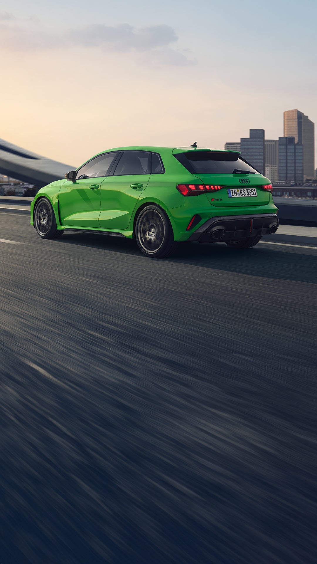 Grüner Audi RS 3 Sportback fährt auf einer Brücke vor städtischer Skyline, Rückansicht.