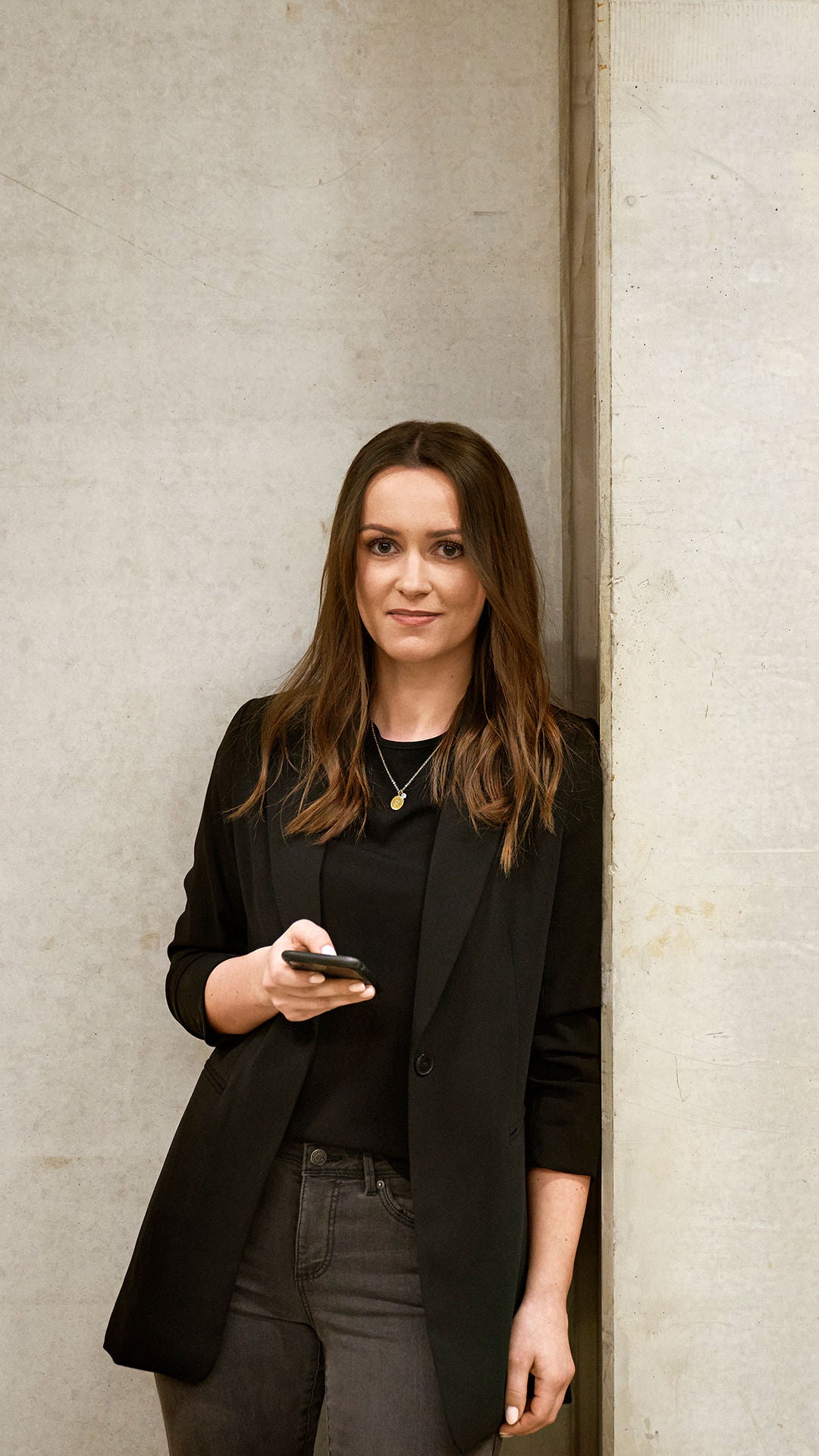 Sabrina is leaning against a wall, looking into the camera and holding a cell phone in her hand