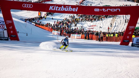 Ein bremsender Skifahrer im Ziel in Kitzbühel. Auf dem roten Zielbogen sind ein Audi-Logo und die Schriftzüge Kitzbühel und e-tron zu sehen.