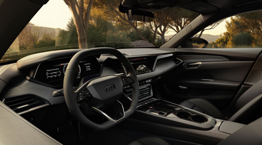Het interieur van een Audi e-tron GT quattro met zicht op een tuin. De zetels, het stuur en het dashboard zijn zichtbaar in het licht van een zonsondergang.