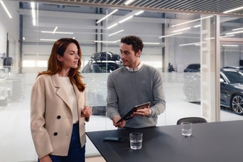 Mann zeigt Frau ein Tablet in modernem Audi-Autohaus, auf dem Tisch davor stehen zwei Gläser Wasser und ein Smartphone.