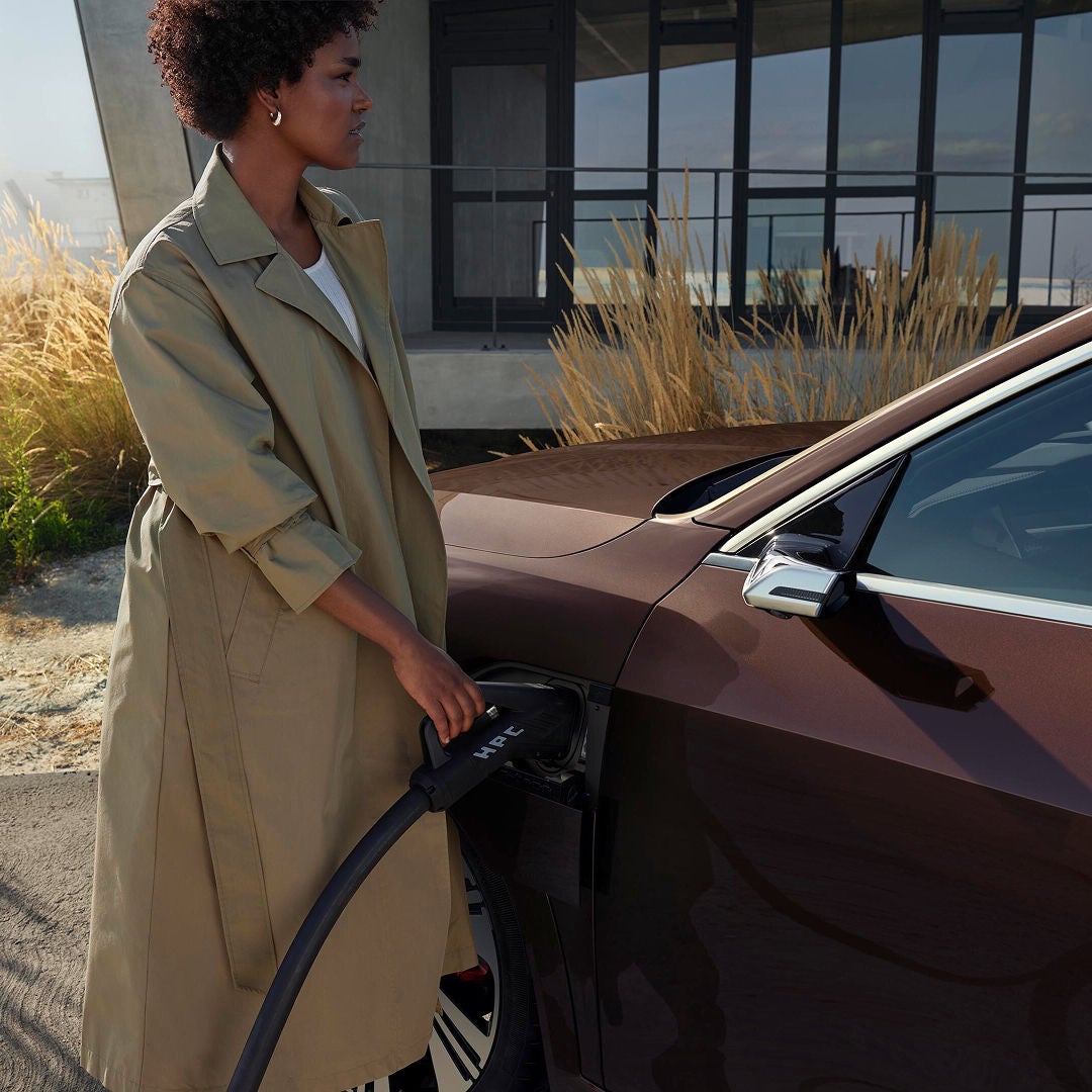 Woman in a beige trench coat charges a brown electric car in a modern setting with tall grass and a building in the background, evoking eco-friendly themes.