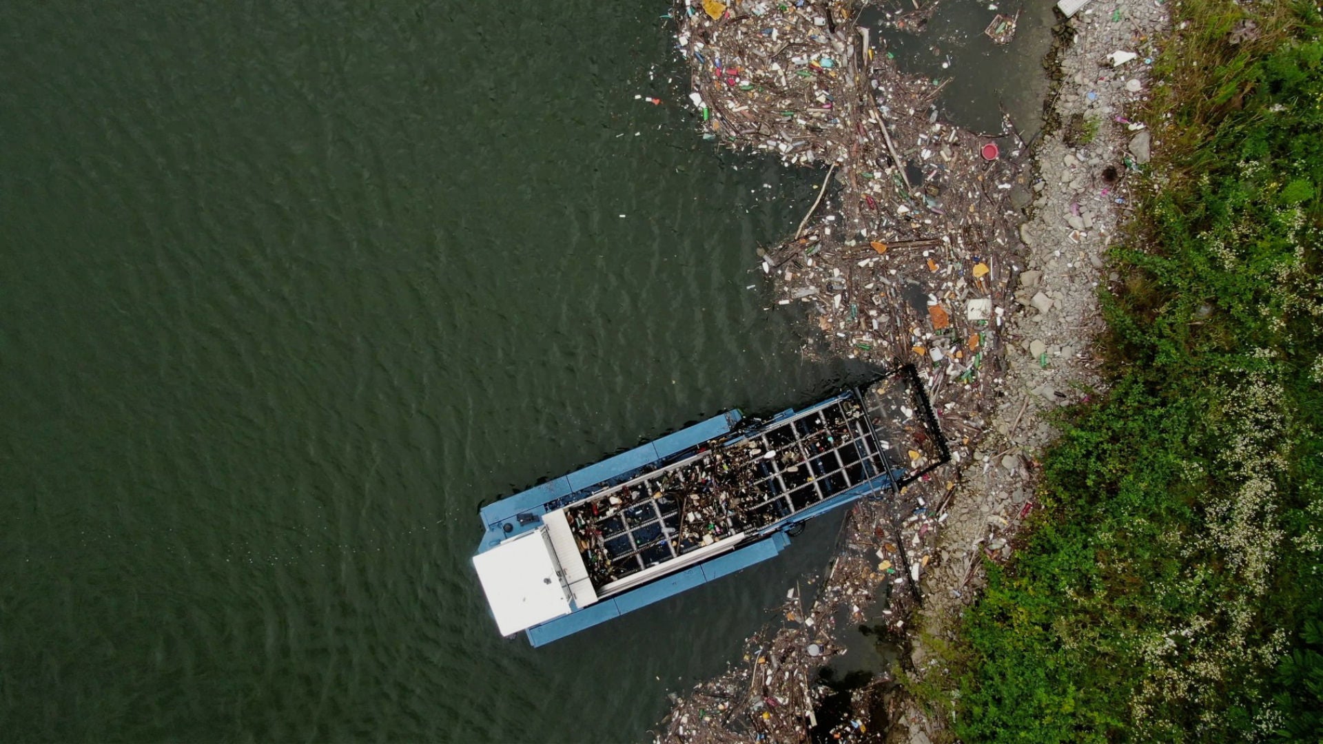 A rubbish collection boat from above on a riverbank surrounded by rubbish.