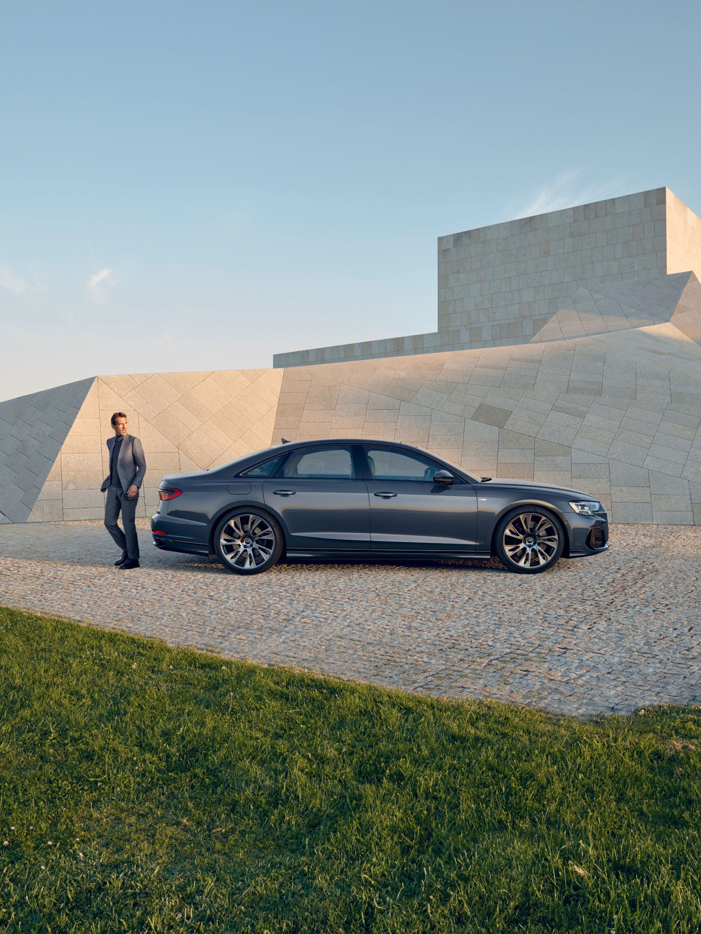 A person in a suit standing next to a sedan on a cobblestone surface with a geometric building backdrop.