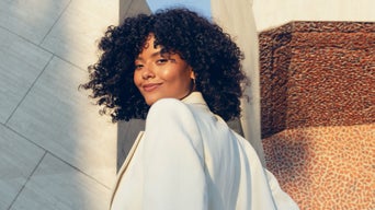 Frau mit lockigem Haar und hellem Blazer lächelt in die Kamera, moderne Architektur im Hintergrund bei Sonnenlicht.