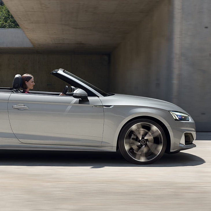A silver convertible car driving under a bridge.
