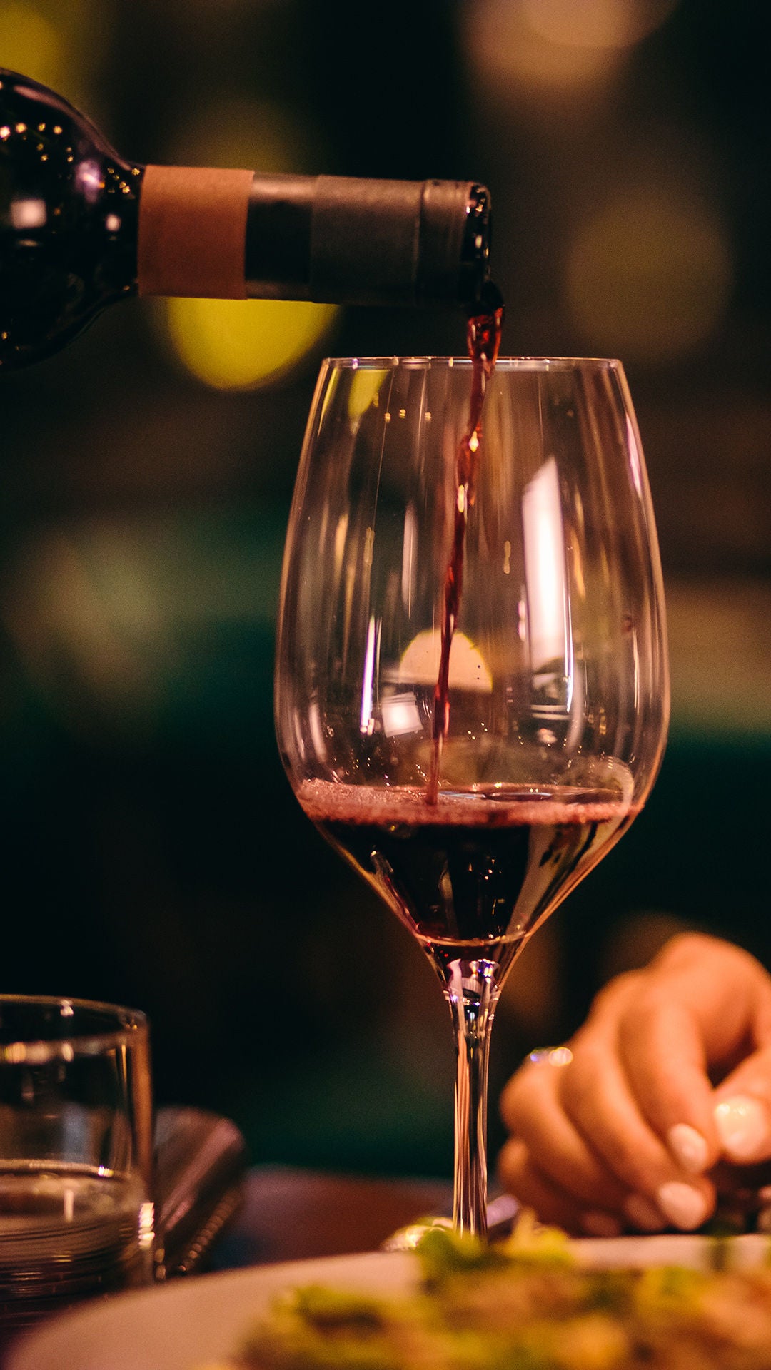 Rotwein wird in ein Weinglas eingeschenkt; im Hintergrund sitzt eine Person an einem warm beleuchteten Esstisch.