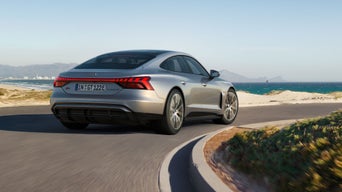 Silberner Audi S e-tron GT fährt an Küstenstraße eine Kurve entlang, Meer und Berge im Hintergrund bei klarem Himmel.