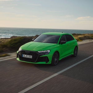 A vibrant green Audi RS 3 Sportback speeds along a coastal road with the ocean in the background under a clear sky.