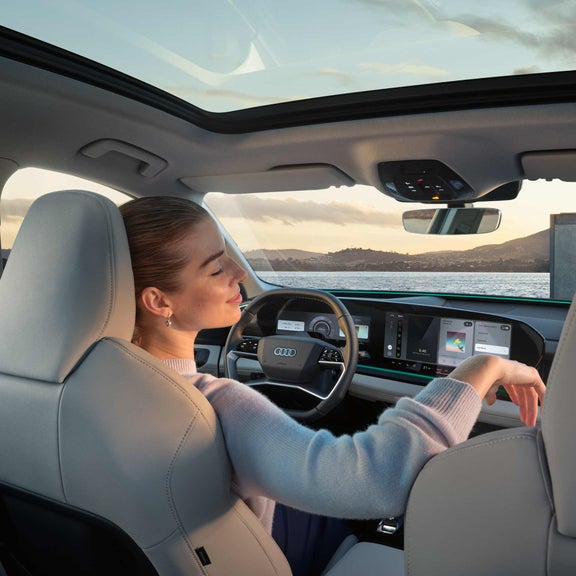 Innenraum des Q6 SUV e-tron aus Rücksitzperspektive. Eine junge Frau sitzt auf dem Fahrersitz und lehnt sich mit geschlossenen Augen zurück. Durch die Windschutzscheibe sieht man das Meer und Berge.