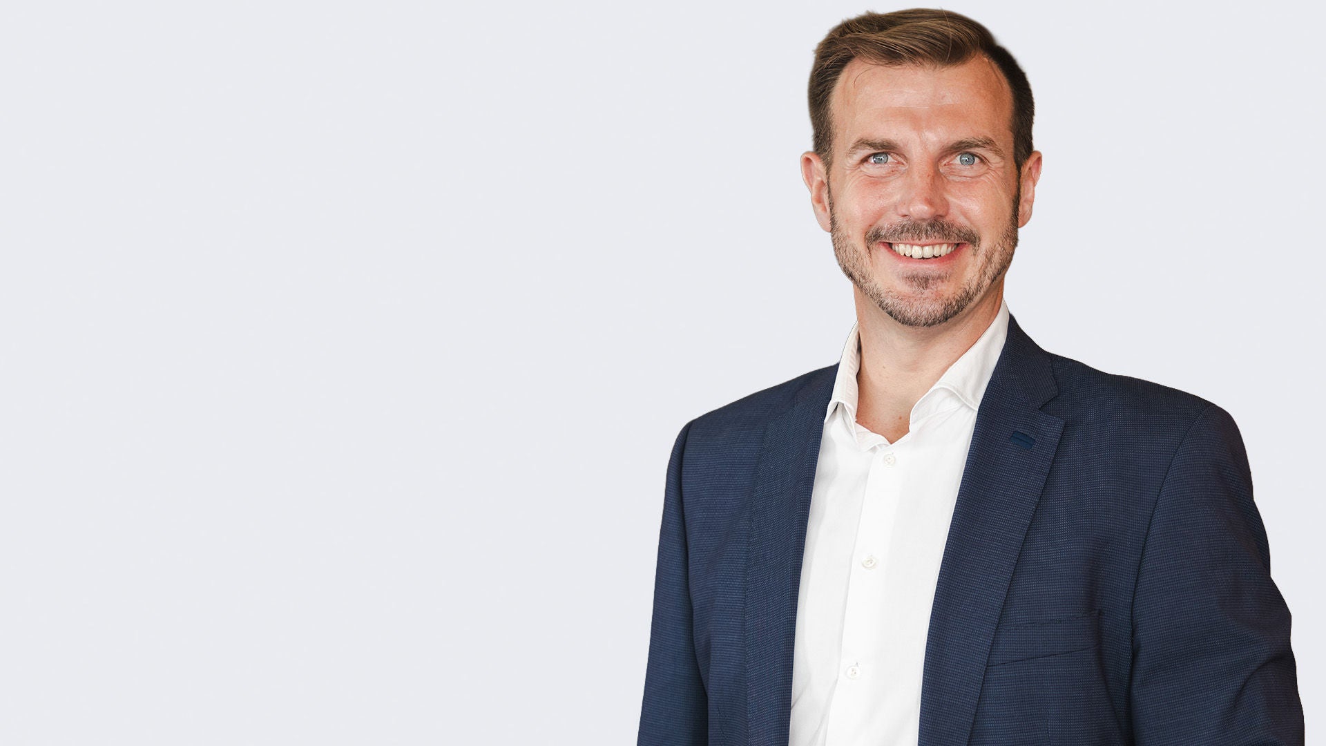 Mann im dunklen Sakko, hellem Hemd und gepflegter Erscheinung vor einem grauen Hintergrund.