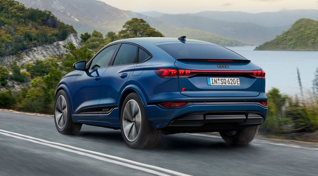 Schräge Heckaufnahme des SQ6 Sportback e-tron, welcher eine Straße an einem See in einer bergigen Landschaft entlangfährt.