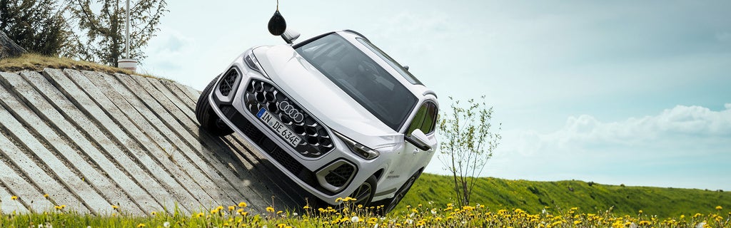 Weißer Audi Q5 SUV e-hybrid auf schräger Betonrampe; Frontansicht mit Audi-Logo, Blumenwiese und Himmel im Hintergrund.