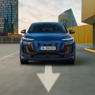 Frontansicht eines blauen Audi SQ6 Sportback e-tron auf Parkdeck mit Pfeilmarkierung, Hochhäuser im Hintergrund.