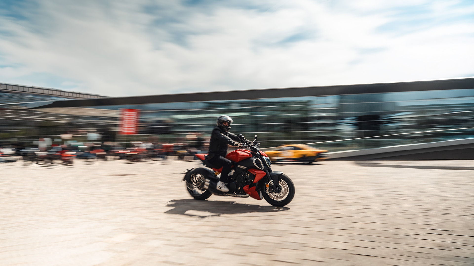 Person mit Helm fährt rotes Motorrad über Platz, modernes Gebäude im Hintergrund.