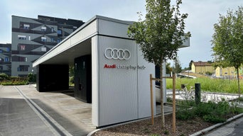 Audi Charging Hub bei Tag mit weißer Fassade, Audi-Logo und Grünflächen im Vordergrund, im Hintergrund Wohngebäude.