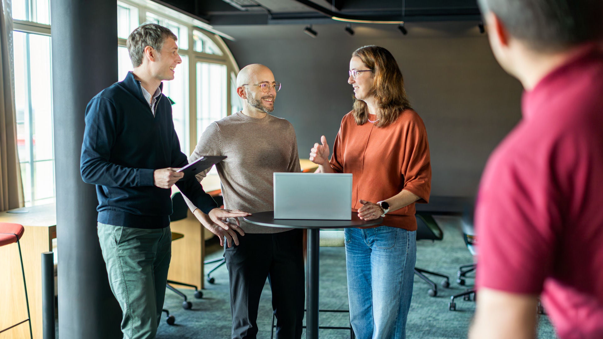 Several people are standing and sitting at a high table with laptops and talking to each other.