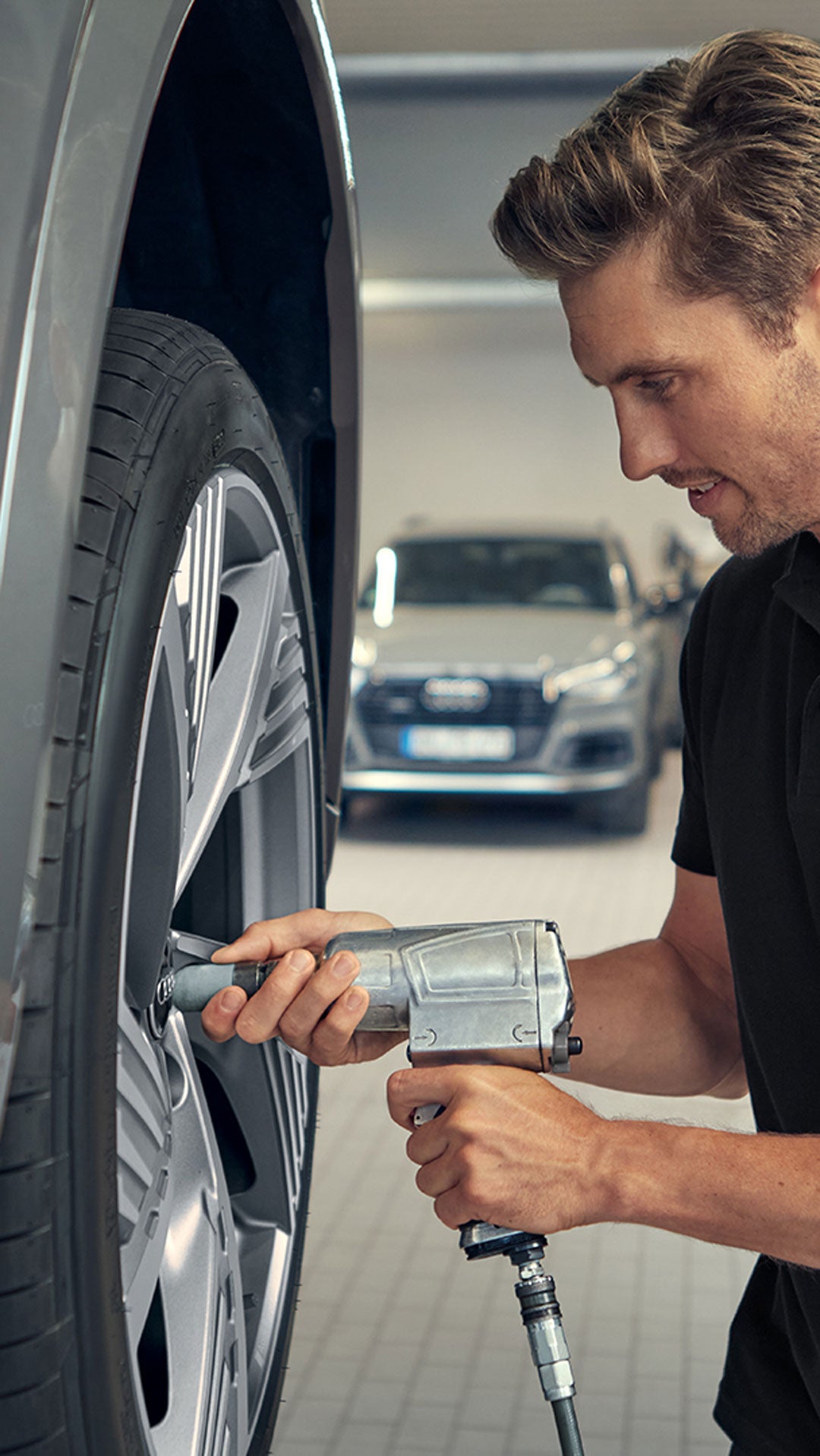 Ein Audi-Mitarbeiter montiert mit einem Druckluftschrauber ein Rad an einem Fahrzeug in einer Werkstattumgebung.