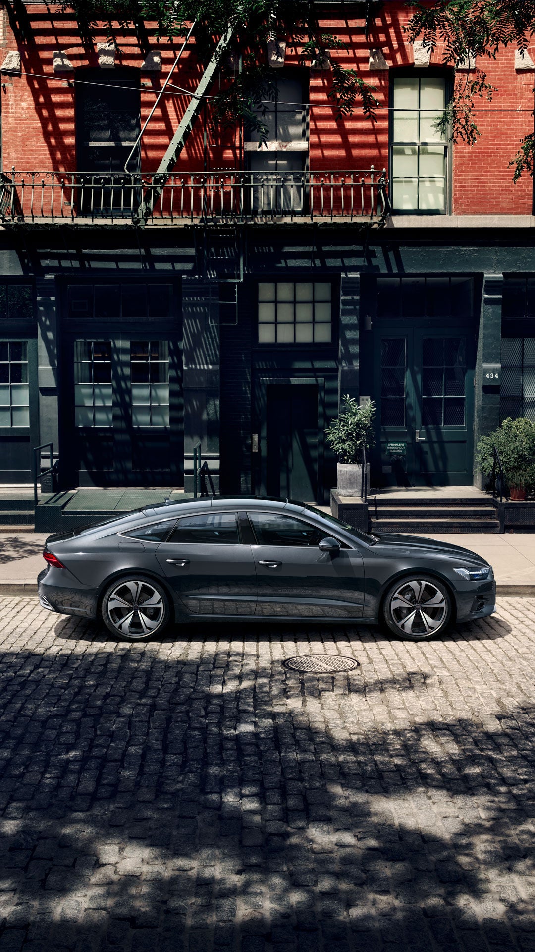 Grauer Audi A7 Sportback parkt seitlich auf Kopfsteinpflaster vor einem Gebäude mit dunkler Fassade und großen Fenstern.